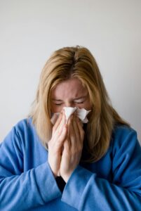 A woman in a blue robe is sneezing into a tissue, illustrating symptoms of a cold or flu indoors.