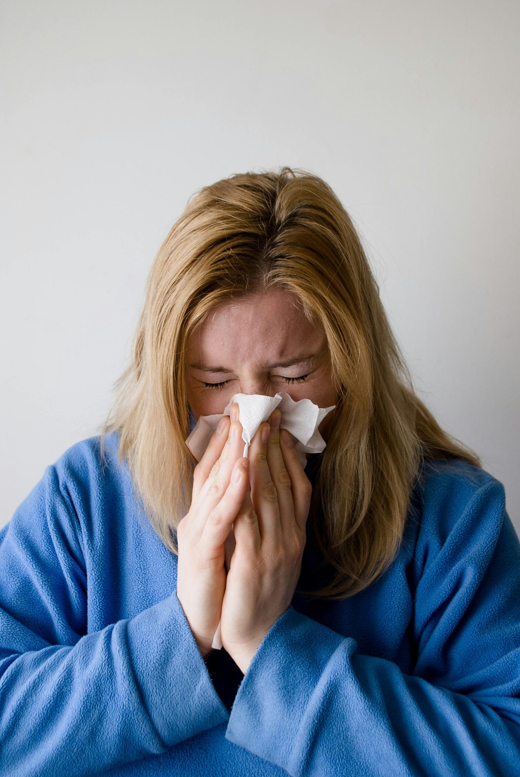 A woman in a blue robe is sneezing into a tissue, illustrating symptoms of a cold or flu indoors.